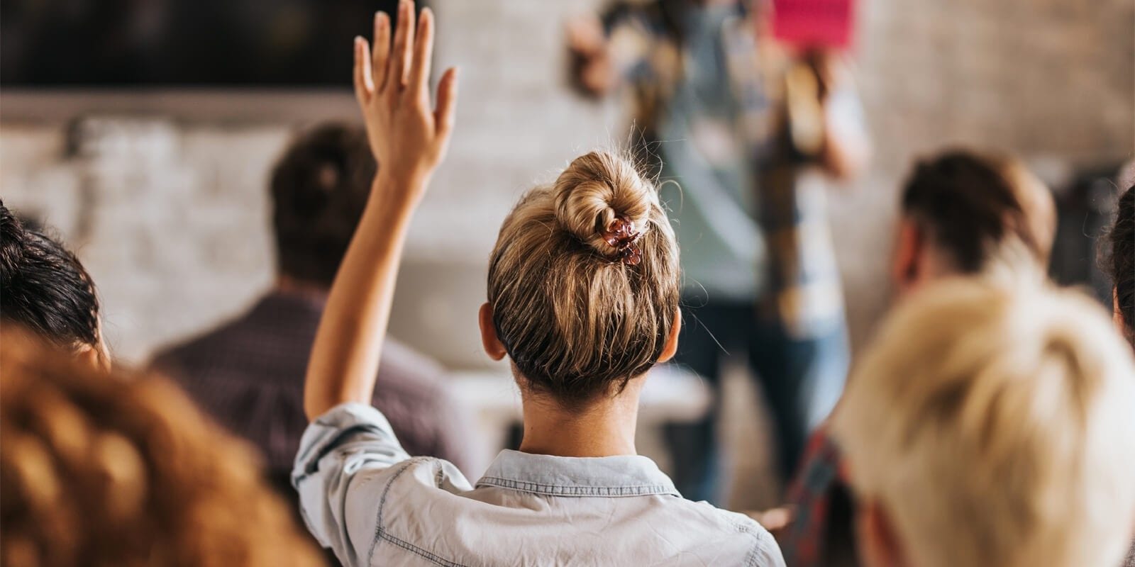 A person with a bun hairstyle raises their hand in a classroom setting, surrounded by other attendees facing forward.
