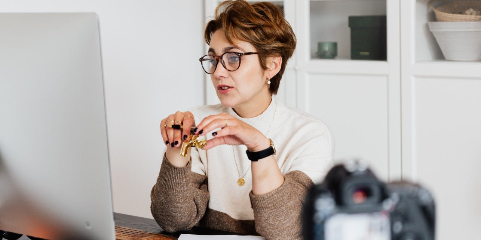 A person sits at a desk, adjusting a watch, with a computer, camera, and white office decor visible.