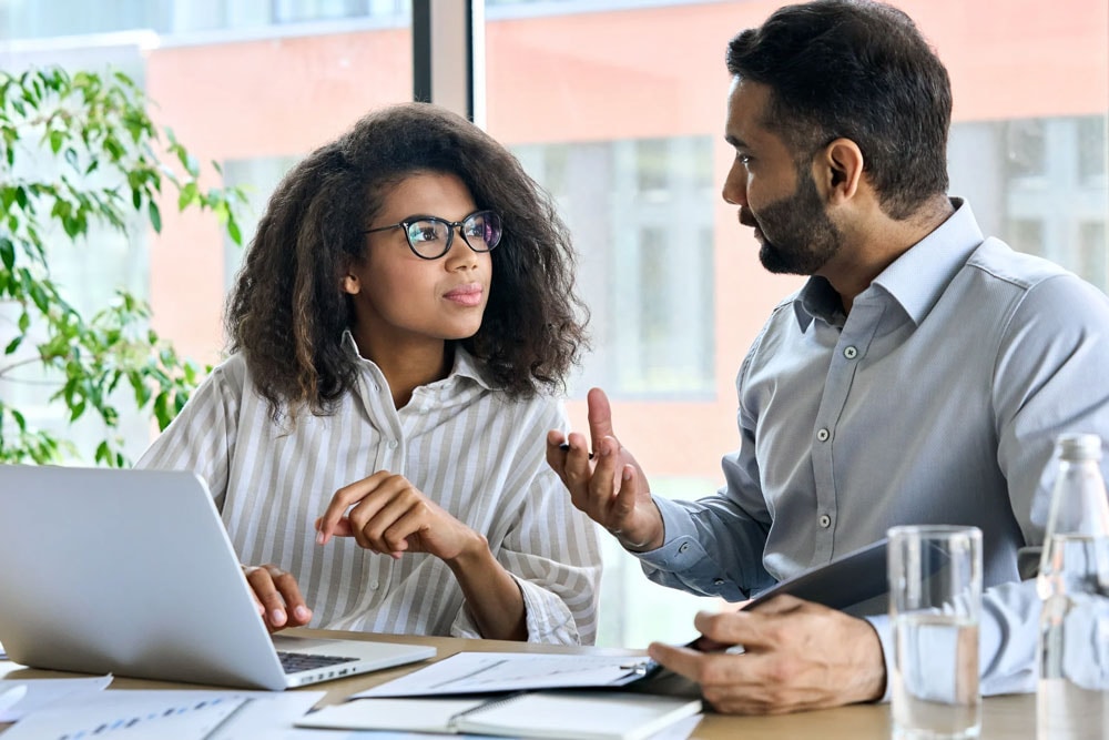 Two professionals, a man and a woman, are engaged in a discussion at a table with a laptop and documents in a bright office setting.